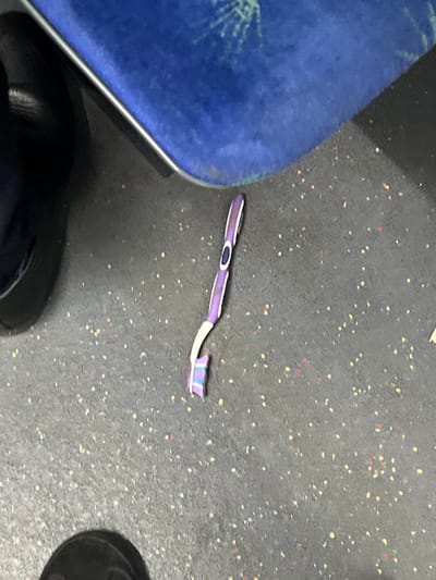 The image depicts a broken electric toothbrush on the floor of a Jerusalem Light Rail car, located in West Jerusalem, Israel. The toothbrush handle is predominantly purple with white accents and features a central blue oval button. The detached brush head, showing blue and white bristles, lies immediately adjacent to the handle. The floor is dark gray with multicolored speckles, including red, yellow, green, and light blue. A blue upholstered seat with a light trim and a subtle patterned fabric is visible in the upper portion of the frame. The toe of a dark-colored shoe is partially visible in the lower-left foreground. No human figures are fully visible, and no actions or interactions with the toothbrush are depicted.