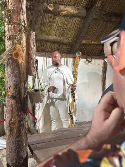 The image captures a scene set within a rustic, open-air structure, possibly an outdoor stage or exhibit, in the city of Indjija Municipality, Serbia. The dominant subject is a bald man with a beard, wearing a white robe and pants, and a necklace with a circular pendant. He is holding a long, ornate staff with a carved animal head at the top, made of wood with decorative bands. The man appears to be standing in front of a white wall with a rough texture, possibly made of mud or plaster, and part of a thatched roof is visible above. To the left of the man, a rough-hewn wooden post stands tall, with a burlap sack hanging from it containing a small plant. A red rope is also visible, attached to the post. In the foreground, partially obscuring the view, is the arm and hand of another person, along with a wooden table. The overall impression is that of a historical reenactment or a cultural display.