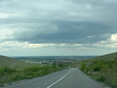 This is an eye-level, full, outdoor shot taken on a paved, one-lane road with a broken white line down the center. The road runs between grassy hills on either side and descends into a flat area in the distance with a small town with red roofs surrounded by agricultural buildings and fields. The sky is cloudy and gray. In the far distance, there appear to be tall, narrow industrial structures. The photo appears to be taken near Isperihovo, Bulgaria, based on the style of buildings and the landscape.