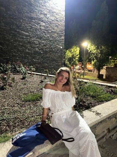The image captures a nighttime scene in Mogadouro, Portugal. A single female subject is seated on a light-colored stone ledge, facing the camera with a slight smile. She has long blonde hair, wears glasses, a gold-colored necklace, small earrings, and a pink bracelet on her right wrist. Her attire consists of a white off-the-shoulder top with eyelet embroidery and a matching long, wide-legged skirt with similar embroidery. Beside her on the ledge rests a dark brown woven handbag and a folded blue garment.
The background to the left features a tall, dark gray, textured stone wall, illuminated from its base, highlighting its rugged surface. Below this wall, a garden bed with dark soil or mulch contains scattered low-lying green and reddish-leafed plants. To the right of the subject, the background shows several green trees, some brightly illuminated, and a prominent streetlight emitting a warm yellow glow. Further in the distance, lights from buildings are visible against the dark night sky, suggesting an urban or town setting.