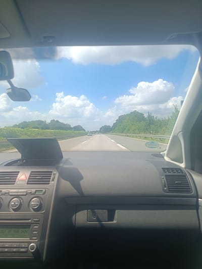 This image captures a view from inside a car driving on a highway. The dashboard of the car is visible in the foreground, with a radio display showing the text "OUT OF THE DARK". The road stretches ahead, with other cars in the distance. The setting is a sunny day with a blue sky and scattered white clouds, indicating pleasant weather. Lush green trees and fields line the sides of the highway. The scene suggests a road trip or travel through a rural or suburban area in Germany, given the location context of Bad Sassendorf. The overall mood is one of travel and openness, with the bright sky and clear road contributing to a sense of forward momentum.