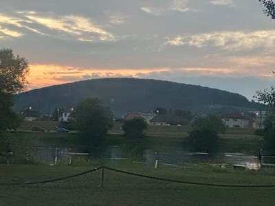 The image depicts a dusky landscape with a body of water in the foreground, a grassy bank, and a town nestled at the base of a large, tree-covered hill. The sky is overcast with streaks of orange and yellow from the setting sun. In the town, several houses with red or white roofs are visible, along with streetlights. Two cars, one red and one blue, are parked on a road. A person is walking on the grass near the water's edge. The scene is set in Zamarovce, Slovakia.