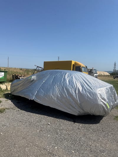 A silver car cover is draped over a car parked on a gravel surface. The car cover is reflective and appears to be made of a metallic material. The car is parked outdoors, with a clear blue sky overhead. In the background, there are two trucks, one yellow and one white. There are also power lines, some trees and a green dumpster. The location is identified as the city of Slanchevo, Bulgaria.