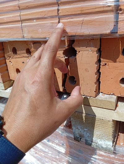 A hand, with skin showing a slight tan and visible veins, is positioned in the foreground, with the index finger pointing towards a stack of bricks. The bricks are reddish-orange and appear to be wrapped in clear plastic sheeting. Some of the bricks show signs of damage, with cracks and chips along their edges and surfaces. The stacking of the bricks is supported by wooden blocks, some of which are also showing weathering and white residue. The scene appears to be outdoors, possibly a construction site or a storage area for building materials, indicated by the presence of bricks and the lighting which suggests daylight. The background is out of focus but shows more of the stacked bricks and plastic wrapping. There is no visible text in the image. The context provided places the scene in Қызылжар ауылдық округі, Kazakhstan.