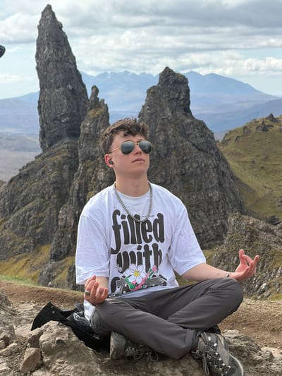 A young man is sitting in a cross-legged position on a rocky outcrop with a mountain range in the background under a cloudy sky. The man is wearing sunglasses, a white t-shirt, grey trousers, and hiking boots. He has a silver chain around his neck. The landscape suggests a mountainous region, possibly a part of the Scottish Highlands, particularly in the Isle of Skye near Portree, United Kingdom.