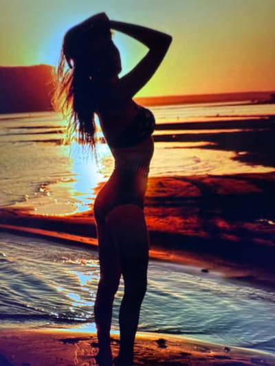 A silhouetted woman in a bikini stands on a beach with her hand on her head, looking out at a sunset over the ocean. The sky is a gradient of yellow, orange, and red. The water reflects the colors of the sky, creating a shimmering effect. The sand is dark and wet from the receding tide. There are no buildings or other structures visible. The woman is positioned in the foreground, facing left. The scene appears to be a natural landscape with no indication of the city of Bevern, Germany.