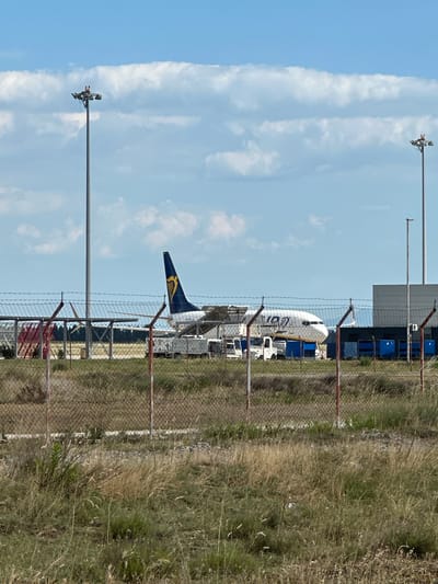 An airplane is parked at an airport, with its tail visible. The airplane has a blue and yellow tail with a logo. In the foreground, there is a chain-link fence with barbed wire on top, separating the viewer from the airplane. Beyond the fence, there is dry grass and vegetation. In the background, there is a building, possibly a hangar or terminal, and more of the airplane. Two tall poles with lights or antennas are also visible. The sky is blue with some white clouds. The scene appears to be outdoors during the day, likely under sunny weather conditions. The image is taken from a low angle, looking upwards towards the airplane. There is no visible text in the image.