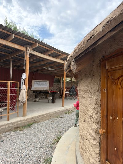 A medium shot captures a scene in Кызыл-Туу, Kyrgyzstan. In the foreground, the textured, mud-plastered wall of a rustic building with a wooden door is visible on the right. A child in a pink shirt and green pants stands with their back to the camera, looking towards a covered outdoor area. This area, likely a workshop or gathering space, features a corrugated metal roof supported by wooden beams and poles. Hanging from a pole are two pieces of clothing. A whiteboard with diagrams is displayed on the back wall of the covered area, and a rough wooden table stands in front of it. The ground is covered with gravel and some sparse green vegetation. In the background, a wooden lattice structure and a black plastic bag are present, with a stone wall and trees visible further back. The sky above is cloudy.