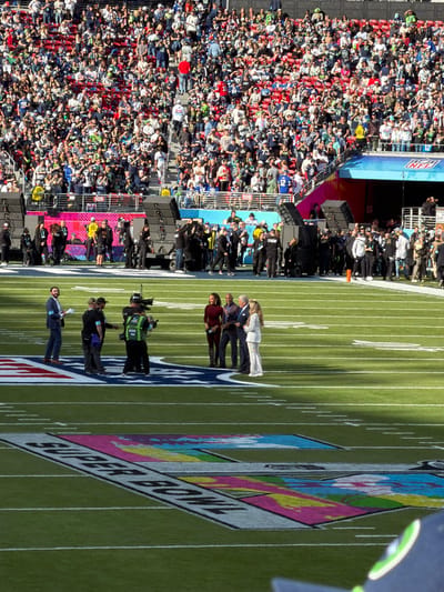 A large crowd of people fills the stands at a stadium. On the field, several people, including a group of four, stand near a large Super Bowl logo. Cameramen and other personnel are positioned nearby. The field has yard lines and markings. The stadium appears to be brightly lit.