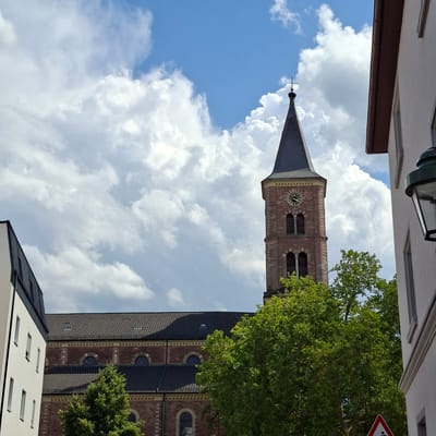 The image shows a view of a church tower with a clock face, partly obscured by trees, with a bright blue sky and white clouds in the background. To the left of the church tower, part of a modern building with white walls and dark metal trim is visible. To the right, a portion of a building with white walls and a dark gutter is present, along with a black street lamp. The church itself is made of red brick with arched windows. The church tower is tall and tapers to a dark spire, with a clock showing approximately 2:52. The setting appears to be an urban environment in Baden-Baden, Germany, based on visual cues such as architectural style and the presence of a street lamp and traffic sign.