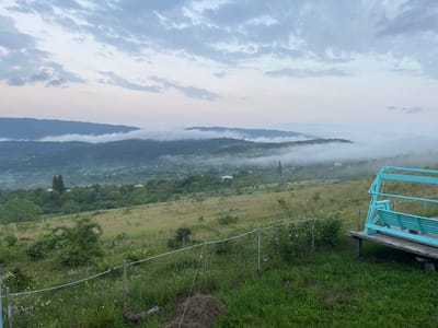 The image depicts a serene landscape at what appears to be either dawn or dusk, characterized by a misty haze obscuring the distant hills and valleys. In the foreground, a vibrant turquoise swing set stands on a wooden platform, inviting relaxation. A simple wire fence separates the immediate grassy area from the undulating terrain beyond, which is dotted with scattered houses and dense greenery. The sky is overcast with soft, diffused light, suggesting a tranquil atmosphere. The location is identified as Nikortsminda, Georgia.