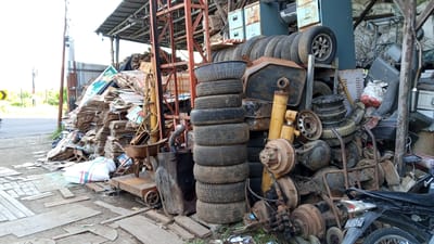 This image depicts a cluttered outdoor storage area filled with discarded materials, likely a junkyard or recycling facility. The scene is dominated by stacks of old tires, piles of flattened cardboard boxes, and various automotive parts like axles, shock absorbers, and fuel tanks. A rusty metal structure, possibly part of a building's framework or a large piece of machinery, stands prominently in the background. To the left, a road with a yellow traffic sign and some greenery suggests the location is near a thoroughfare in a semi-rural or industrial area. The ground is covered in dirt, debris, and scattered wooden planks.
The overall impression is one of disarray and abandonment, with items piled haphazardly. The lighting suggests it is daytime, with bright overcast skies, as there are no strong shadows. The weather appears to be dry. No people are visible in the frame.
Notable details include a white sack with colored stripes lying on the ground, a wheelbarrow, and the partial view of a black motorcycle with a visible license plate. The license plate displays the text "BL 8816 ZS". The setting appears to be Cotbadatunong, Indonesia, based on the location context provided. The atmosphere is utilitarian and industrial, with no emotional cues conveyed.