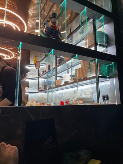 This image, captured in Basiglio, Italy, presents an interior view of what appears to be a modern restaurant or bar, likely during the evening due to the artificial lighting. The focal point is a tall, multi-tiered display unit featuring dark, marble-patterned vertical supports and clear glass shelves illuminated with a subtle green glow along their edges.

The glass shelves are meticulously arranged with various dishware, including stacks of green and white plates, bowls, and drinking glasses. Decorative elements include a colorful Nutcracker-like figure on one shelf and a bottle, possibly of wine. Lower shelves hold small red and yellow condiment bottles. Reflections of the interior lighting and possibly a person can be seen on the glass surfaces.

To the right of the display, a dark, marble-patterned tabletop is visible. A black menu, featuring the text "KYOHO RESTAURANT" and a chopstick logo, is placed on the table. A person's arm and a partial view of their head/shoulder, wearing a dark t-shirt, are visible in the lower right, suggesting they are seated at the table. Another hand is also visible near the menu. Curved, warm-toned strip lights are reflected on the left side, enhancing the sophisticated and contemporary atmosphere of the setting.