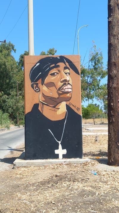 The image shows a mural of Tupac Shakur painted on a concrete pillar in the city of Lambi, Greece. The mural depicts Tupac from the chest up, wearing a black t-shirt and a bandana. He has a gold chain around his neck with a white cross pendant. The background of the mural is a solid brown color, and the artist's signature "ScOotH_88" is visible on the right side of the portrait.

The pillar is located outdoors, next to a road, with trees and a clear blue sky in the background. A wooden utility pole stands to the right of the pillar. The ground around the pillar is covered with dry leaves and dirt. The overall scene is a public space, possibly a street or park area.