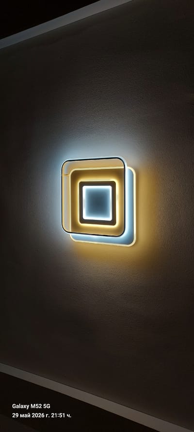 A ceiling-mounted light fixture is the central focus of the image. The fixture is a square design with concentric squares of white and yellow light. The surrounding ceiling is off-white with a textured surface. There is a black border visible on the left and right edges of the image. The date and time of the photo, "29 май 2026 г. 21:51 ч.", is embedded at the bottom right corner. The photo was taken with a Galaxy M52 5G. There is no evidence of people or the city of Canals, Spain.