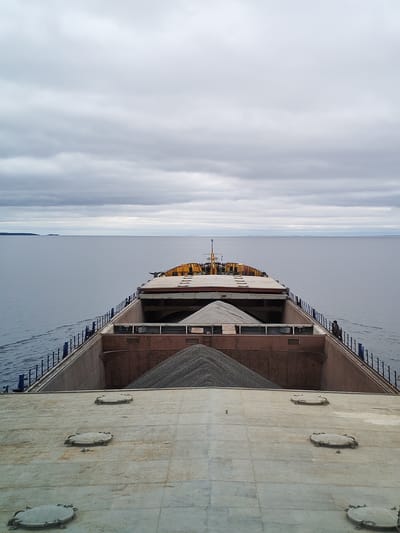 This image provides an elevated, forward-facing perspective from a bulk carrier or cargo ship navigating a vast, calm body of water. The ship's open cargo holds are prominent, filled with large, distinct mounds of gray gravel or aggregate, suggesting the vessel is transporting construction materials. In the foreground, several round, latched hatch covers are visible on the deck.

On the ship's starboard side, near the railing, a lone individual can be seen observing the surroundings, adding a human element and scale to the scene. The vessel is actively underway, indicated by the subtle wake trailing behind it on the smooth water surface.

The sky is uniformly overcast with thick, gray clouds, casting a soft, diffuse light over the entire landscape. A distant, low-lying landmass is faintly visible on the left horizon. The expansive water and flat horizon suggest a large lake or sea. Given the context of Medvezhyegorsk, Russia, this scene likely depicts the ship's journey on a large inland water body such as Lake Onega. The overall atmosphere is calm and functional. No visible text is present in the image.