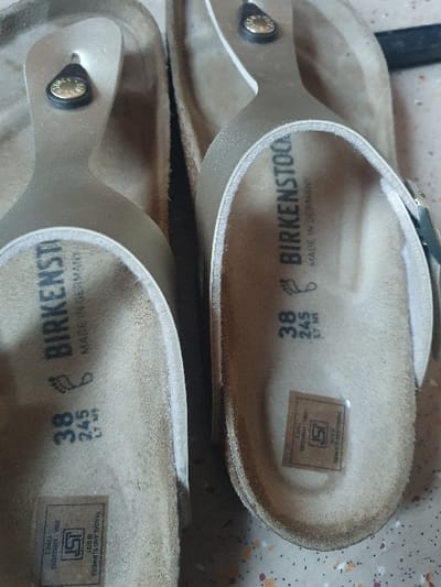 The image displays a pair of gold-colored Birkenstock thong-style sandals positioned on a speckled, multi-toned floor, located in Sadulpur, India. Both sandals feature a tan suede footbed and metallic gold-toned straps. The footbeds are visibly stamped with "BIRKENSTOCK" and "MADE IN GERMANY," along with the size "38" and "245" mm. Each footbed also bears a rectangular sticker displaying the "ISI" (Indian Standards Institution) mark, indicating "SANDAL AND SLIPPERS IS 6721/2021." A small, dark metallic rivet with a partial "BIRK" inscription is visible on the strap near the toe post of both sandals. The right sandal is in sharper focus and shows a portion of an adjustable buckle on its side strap. The sandals are oriented with their heels towards the top of the frame, presented from an overhead perspective. No individuals are present in the scene, and no specific actions or interactions are depicted. The floor surface beneath the sandals is consistent with a terrazzo or tiled pattern, exhibiting flecks of orange, white, and dark brown against a lighter grey base.