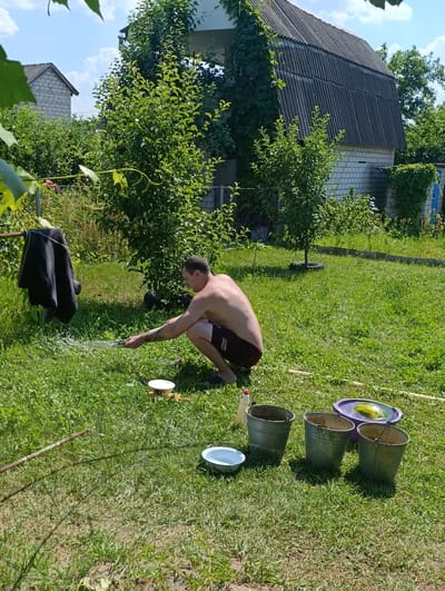 In a sun-drenched, rural backyard in Ladyzhyns'ki Khutory, Ukraine, a shirtless man is crouched on lush green grass, holding a hose and spraying water. The man, who appears to be in his 30s, is wearing dark shorts and has a tattoo on his arm. He is focused on his task, directing a stream of water across the lawn.
In the foreground, several objects are scattered on the grass: three metal buckets, a white bowl, a plastic purple basin containing a watermelon, and a bottle of what appears to be cleaning liquid. A black garment hangs from a clothesline to the left of the frame.
The background features a residential scene with two buildings. The structure closest to the viewer has a grey, corrugated roof and white brick walls, partially covered in ivy. Behind it, another building is visible, also constructed with white bricks. Abundant green trees and bushes fill the yard, creating a verdant and somewhat overgrown atmosphere. The sky above is blue with scattered white clouds, suggesting a clear and warm day. The overall impression is one of a casual, outdoor chore taking place in a rustic, possibly summer setting. There is no visible text in the image.