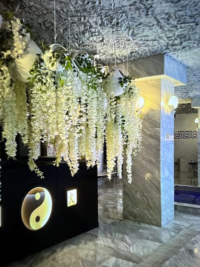 The image depicts an interior scene, possibly a spa or a reception area, with a dominant gray textured wall on the left and a marble-like finish on the upper structure. A large arrangement of white flowers cascades downwards, adding a soft, organic element to the otherwise modern and somewhat stark environment. The flowers are densely packed, creating a visually striking display.
In the background, a dark wall features a prominent illuminated Yin and Yang symbol, a classic representation of balance and harmony. Above it, a smaller square illuminated panel displays a symbol that appears to be a stylized letter or character. The lighting in the scene is soft and warm, with some direct illumination on the flowers and decorative elements, suggesting it is not daytime.
The arrangement of flowers suggests a decorative purpose, perhaps to create a welcoming or calming atmosphere. The overall impression is one of modern design with a touch of natural beauty and spiritual symbolism. The setting appears to be indoors, with no visible windows or outdoor elements. The location context provided (Denver, United States) does not offer specific visual cues within the image itself. There is no discernible activity or event taking place, implying the image captures a static, staged environment.