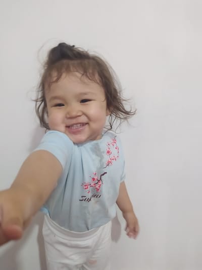 This is a portrait-oriented, horizontally oriented, medium shot of a young child lying on a white surface, possibly a bed or floor. The child, appearing to be a toddler, has dark, wavy hair and a fair complexion. They are looking towards the camera with a wide, happy smile, showing visible teeth. The child's eyes are dark and appear to be looking slightly to the left of the camera.
The child is wearing a light blue t-shirt with a pink cherry blossom design and the word "Sakura" written in dark lettering. Below the t-shirt, white fabric is visible, suggesting they are also wearing white shorts or pants. The child's right arm is extended towards the camera, with the hand blurred due to motion.
The background is a plain, light-colored wall, creating a simple and uncluttered setting. The lighting appears to be soft, possibly natural light from a window, casting subtle shadows on the white surface. The overall mood is joyful and innocent, conveyed by the child's bright smile. There are no clear indicators of the time of day, weather, or specific location beyond the provided context.