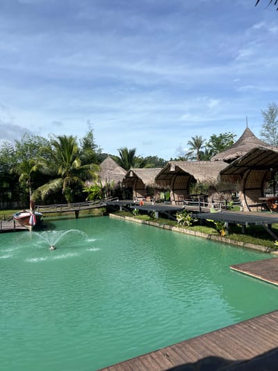 The image depicts a serene resort scene in Tambon Pa Kae Bo Hin, Thailand. The setting features traditional thatched-roof bungalows nestled amongst lush tropical foliage, including palm trees. A vibrant, turquoise swimming pool or lagoon is a prominent element, with a wooden deck bordering one side. The pool is enhanced by a fountain, creating a dynamic visual with water sprays.
In the foreground, a wooden jetty extends into the water, adorned with colorful decorative elements that resemble a pinwheel. A person is visible near the end of the jetty, possibly preparing to enter the water or enjoying the view. The sky above is a clear blue, punctuated by scattered white clouds, suggesting a pleasant, sunny day, likely in the late morning or early afternoon.
The overall impression is one of a tranquil and inviting tropical getaway. The architecture of the bungalows, with their natural materials and conical roofs, speaks to a local style, reinforcing the Thai location. The calm water and the presence of greenery contribute to a sense of relaxation and natural beauty. There is no visible text in the image.