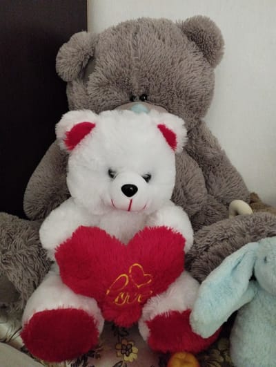 A white teddy bear with red accents on its ears and paws is prominently positioned in the foreground, holding a red heart with "love" embroidered in yellow. Behind it sits a larger, gray teddy bear. To the right, a light blue stuffed rabbit with long ears is partially visible. The stuffed animals are arranged on a surface with a floral pattern. The background consists of a dark vertical surface on the left and a plain white surface on the right. The scene appears to be indoors. There is no discernible city or location information.