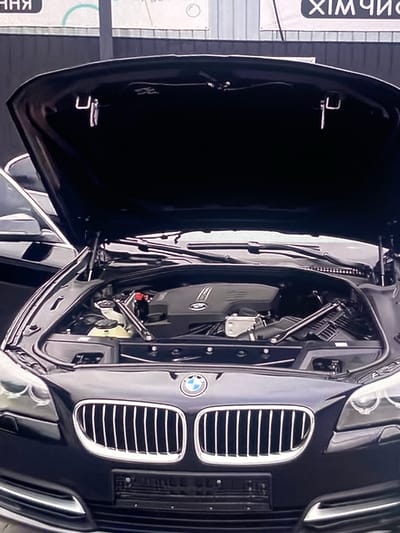 An exterior, eye-level, medium shot shows the open hood of a dark-colored BMW sedan. The engine and various car parts are visible under the hood. The front grill and headlights of the car are also visible. The background appears to be a building with banners that have text in Cyrillic alphabet. The scene is located on a street in Rokytne, Ukraine.