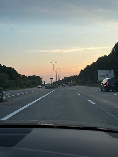 The image depicts a view from a car traveling on a highway during dusk. The sky is a gradient of pale blue and pink, with wispy clouds. On the right side of the highway, there is a sign with directional information in Russian, indicating directions to "KAZAN" and other locations. A black minivan with license plate "Y592VC716" is visible in the right lane, moving in the same direction as the viewer's car. Other vehicles are present on the highway, and streetlights line the road. The landscape on either side of the highway consists of dense green trees. The foreground shows the dashboard and windshield of the vehicle from which the photo was taken. The location is identified as the city of Zvanka, Russia.