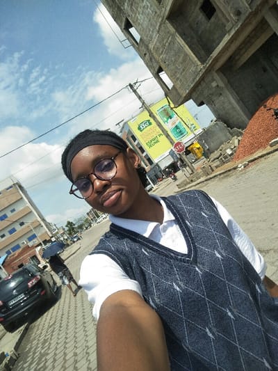 A selfie captures a dark-skinned individual in Cotonou, Benin, on a bright, partly cloudy day. The subject, wearing a black headband, round glasses, a white polo shirt, and a dark blue argyle sweater vest, occupies the foreground, looking directly at the camera with a neutral expression. Sunlight illuminates the right side of the subject's face.

The urban background to the left features a black Mazda CX-5 SUV (license plate BV 8074 AB) parked on a paver-tiled sidewalk. A person holding an open blue umbrella walks away in the mid-distance, with additional buildings and a street visible further beyond.

To the right, an unfinished multi-story concrete building with exposed structural elements is under construction, accompanied by a pile of reddish-brown aggregate material. Two prominent yellow billboards advertise "33cl NOUVEAU FORMAT POUR UN ESPRIT PLUS JEUNE" in French, featuring beverage cans. A red hexagonal STOP sign is mounted below the billboards. Overhead utility wires are visible against the blue sky.