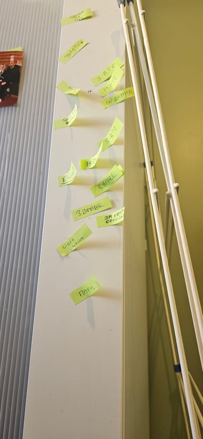 An upward-angled shot reveals a collection of yellow sticky notes affixed to a light-colored horizontal surface, possibly a shelf or a beam. Above this surface, several white metal rods of a drying rack are visible, casting shadows and partially obscuring some of the notes. Below the main surface, a textured gray panel, possibly a radiator or a blind, is seen, and a corner of a framed picture with people is visible on the far left. The wall above the shelf is a pale yellow-green.
The sticky notes are handwritten and arranged haphazardly, some curling at the edges, others overlapping. They display a mix of English and Cyrillic script.
Visible text includes:
- "158" (partially hidden)
- "Щастие" (Cyrillic for "Happiness")
- "4 MEH"
- "3-BTC" (appearing twice)
- "MINED"
- "ON GAMMA"
- "LIBRA VENUS"
- "3 GRADE" (appearing multiple times; some OCR identifies "S&PABE", "39PABE", or "3 драве," suggesting variations in handwriting or Cyrillic characters)
- "3A MOET"
- "CEMETG" (OCR suggests "CEMEGS")
- "Пари" (Cyrillic for "Money")
The mixture of terms like "BTC" (Bitcoin), "MINED," "LIBRA VENUS," and the Cyrillic words suggests a personal collection of notes with diverse themes, possibly relating to cryptocurrency, astrology, and general life concepts. The image is an indoor shot, with no discernible time of day or weather. According to the provided context, the location is Norwich, United Kingdom.