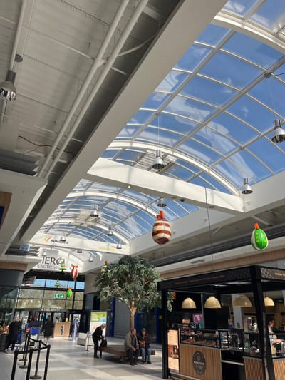 The interior of a shopping mall in Saint-Brice-Courcelles, France, featuring a large, curved glass skylight. The skylight is filled with blue sky and white clouds, and is supported by numerous metal beams. Several hanging light fixtures are suspended from the skylight. Several decorative Easter eggs are hanging from the ceiling. The mall's floor is tiled, and there are various shops and businesses along the perimeter. There are several people walking and shopping, including one person sitting on a bench. There is a large tree in the center of the mall. The image was likely taken in the late afternoon or early evening, as the sun is shining brightly through the skylight. The time is estimated to be around 4 pm in the afternoon. The temperature is estimated to be around 60 degrees Fahrenheit (15.5 degrees Celsius).