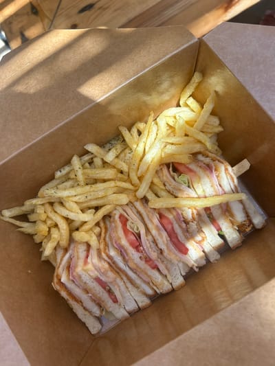 A close-up, overhead shot shows a takeout box filled with food. The box is made of brown cardboard and is open. Inside, a club sandwich is cut into four portions, revealing layers of white bread, ham, cheese, lettuce, and tomato. French fries are piled on top of and to the side of the sandwich. The food is illuminated by sunlight casting shadows across the box and its contents. The setting appears to be outdoors, possibly a table at a restaurant or cafe in Piso Livadi, Greece.