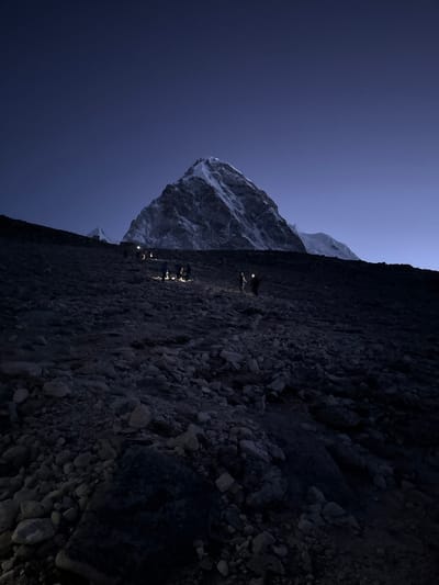 A vertical shot, rotated 90 degrees counter-clockwise from its likely original landscape orientation, captures a group of trekkers ascending a steep, rocky mountain path in the pre-dawn hours or at night. The scene is dominated by dark, muted tones of blue, gray, and black, illuminated primarily by the headlamps of the individuals and faint ambient light.
In the foreground, a rugged, dark gray and black path strewn with various sizes of rocks occupies the lower right portion of the image (when viewed upright). Along this path, several figures wearing headlamps are visible. At least three distinct figures can be clearly seen, their headlamps casting bright, localized pools of light onto the ground directly in front of them, highlighting parts of their bodies and equipment. They appear to be moving upwards, suggesting a strenuous climb.
To the left of the path, a massive, snow-capped mountain or glacier face rises steeply, its contours partially visible through the low light. This majestic peak appears somewhat ghostly, reflecting what little light is available, perhaps from the moon or the very first hints of an impending sunrise. The sky above is a deep, ethereal blue, transitioning from a slightly lighter hue near the horizon to a darker indigo overhead. Tiny pinpricks of light, possibly stars, are faintly discernible in the dark sky, suggesting clear weather conditions.
The overall atmosphere is one of serene solitude and determined effort in a challenging, high-altitude environment. The artificial lights of the headlamps create a strong contrast against the vast, dark landscape, emphasizing the human presence within the grandeur of nature. The image conveys the quiet intensity of an early morning or late-night mountain ascent, likely in the Himalayas, given the context of Khumjung, Nepal. There is no visible text in the image.