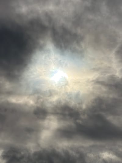 This image depicts a cloudy sky with the sun breaking through in the center, creating a bright, diffused glow. The clouds are varied in texture and shade, ranging from dark, heavy masses to lighter, more wispy formations. There is a small, dark silhouette of what appears to be a bird in the lower left quadrant of the image. The scene is devoid of any specific landmarks that would definitively place it in Gloucester, United Kingdom.