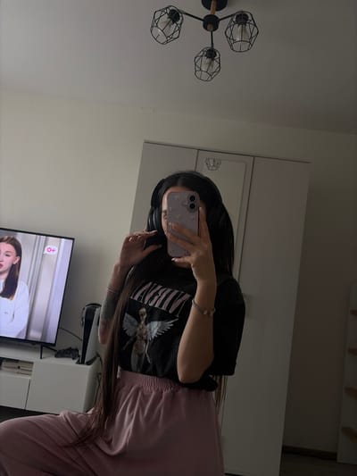 The image shows a person with long dark hair taking a selfie in a mirror. The person is wearing headphones, a black t-shirt with a graphic design, and pink pants. They are holding a light purple phone with a floral design on the case. The background includes a white wall, a light fixture, a partially visible TV screen showing a person's face, and a white cabinet with a gaming console behind the person. There is no information to determine the image was taken in Yoksolovo, Russia.