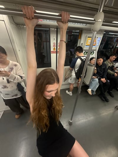 The image shows the interior of a subway car in Changning District, China. Several people are visible, some seated and others lying down on the floor or seats. In the foreground, a young woman with long, reddish-brown hair is holding onto a vertical pole with both hands. She is wearing a black dress and looking towards the right side of the frame with a slight smile. Her arms are extended, showcasing her lean physique.
Behind her, several other passengers are seen resting. One man in a blue and white shirt and white shorts is standing, holding onto a horizontal pole. Further back, three men appear to be asleep or resting on the seats and floor. One man in a green jacket is lying on the seats, while another in a black jacket is also resting. A third man in a blue shirt and jeans is lying down with his head resting on a seat.
The subway car appears to be moderately crowded, with passengers attempting to rest during their commute. The lighting suggests it is daytime, but there are no clear external cues for time or weather. The overall mood seems relaxed, with people taking advantage of the quiet time to rest.
Visible text on the subway car includes signs with Chinese characters and symbols, possibly indicating safety instructions or station information. There are also illuminated display screens with text and graphics, typical of public transportation. The floor of the subway car is gray and shows some scuff marks, indicating regular use.