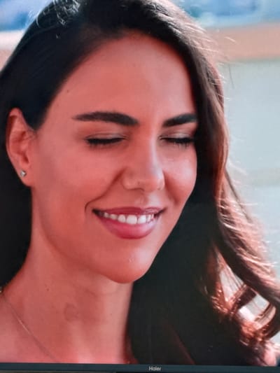 The image is a close-up, waist-up shot of a young woman with dark hair, smiling with her eyes closed. She is wearing a light-colored necklace and a small, silver stud earring in her left ear. The lighting is warm, casting a reddish hue on her skin. Her lips are parted, revealing white teeth, and she has well-defined eyebrows. The background is out of focus, appearing as a blurry wash of light blue and beige, possibly indicating an outdoor setting with natural light. There is a faint, darker mark on her neck. The bottom of the image shows a black bar with the word "Haier" in white text, indicating the image is displayed on a Haier monitor. The location of Uyutnoye, Ukraine, is not depicted or suggested by any elements within the image.