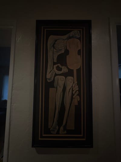 An interior photograph depicts a vertically oriented, framed piece of artwork centrally mounted on a light-colored, textured wall within a residential space in Miami Shores, United States of America. The artwork, characterized by a dark background, features a stylized, elongated, and somewhat gaunt human figure rendered in light grey and beige tones. The figure is depicted in a seated or hunched posture, with its head resting against a light brown, stringed instrument resembling a cello or violin. One hand is positioned near the neck of the instrument, while the other hand, at the bottom right, appears to be gripping the lower section of the instrument. The artwork is enclosed within a thick, dark brown or black frame with a prominent gold or brass inner trim. The scene is dimly lit by an overhead light source. To the left of the artwork, a partial view of a doorway reveals a deeper, darker room with a vertical light source visible on the far wall. To the right, another doorway shows a dark interior. No individuals are visible in the scene.