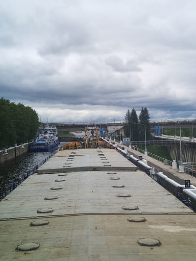 The image shows a barge or large boat navigating a river lock, possibly within or near Rybinsk, Russia. The barge is filled with hatches, and there's a tugboat located on the left side of the barge within the lock. A bridge and other elements of the lock infrastructure are visible in the background. The sky is overcast with dark clouds. The photo appears to be taken from a high vantage point, possibly on the barge itself.