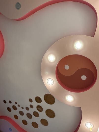 This image shows a close-up, upward-facing view of a decorative ceiling with embedded lights and a yin-yang symbol. The ceiling is predominantly white and pink, with curved sections and circular cutouts. The yin-yang symbol is in shades of brown and white and is centrally located on a circular pink protrusion. Multiple recessed lights illuminate the ceiling. The location is identified as a residential interior in Kandry, Russia. There are no people visible in the image.