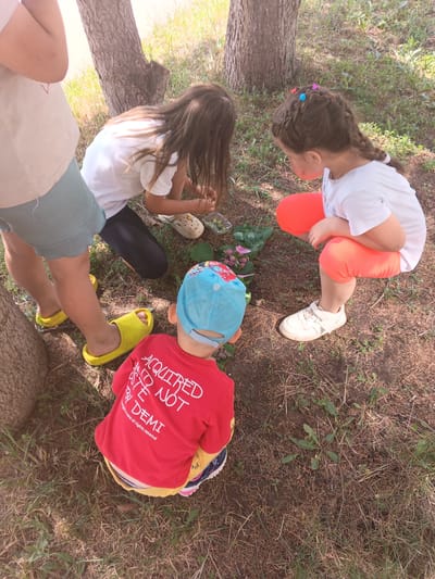 In the city of Tolbazy, Russia, four children are gathered on the ground, engaged in an activity outdoors. The scene, characterized by a natural outdoor environment with trees and dry grass, suggests a park or a wooded area. A young child, seated and facing away from the camera, wears a red long-sleeved shirt with white lettering and a blue baseball cap. To the right, another child, also facing away, is partially visible, wearing a white t-shirt and orange shorts, with braided hair adorned with colorful clips. Two other children are also present, one on the left wearing light-colored shorts and yellow slides, and another in the background in dark leggings and a white t-shirt, kneeling and interacting with items on the ground. In the center, a collection of leaves and possibly flowers are laid out, indicating a nature-related activity. The overall ambiance is one of childhood play and exploration in a natural setting.
