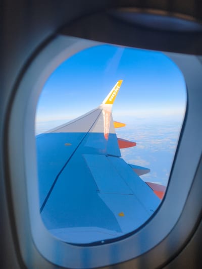 The image is an aerial view through an airplane window, focusing on the wing of an EasyJet aircraft. The wing, predominantly blue with orange winglets, stretches across the frame, with the text "easyJet" visible on the orange tip. Below the wing, a patchwork of land with fields and scattered structures is visible, suggesting a rural or semi-rural landscape. The sky is a clear, bright blue, with faint clouds on the horizon. The curvature of the airplane window frames the scene. Given the context of EasyJet flights and the typical European flight paths, it is plausible that this view is over or near Schwarzheide, Germany, a town located in the Lusatia region of Brandenburg.