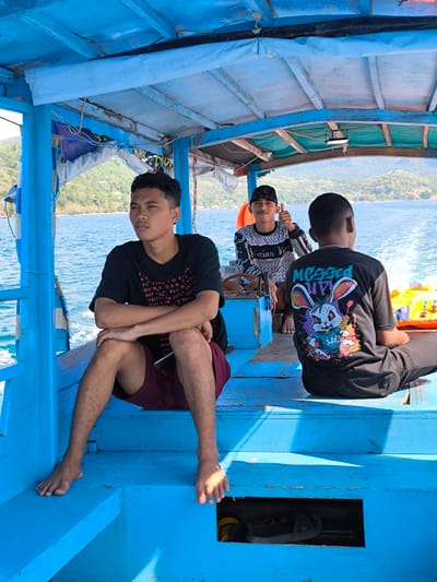 Three young men are seated on a bright blue boat, likely on a journey. The man in the foreground, on the left, wears a black t-shirt with white lettering and dark red shorts. He is looking off to the side with his arms crossed. Behind him, in the center, another young man in a black and white patterned long-sleeved shirt and a black cap gives a thumbs-up to the camera. He appears to be smiling. The third man, on the right, faces away from the camera, wearing a black t-shirt with a colorful graphic of a rabbit and the words "Megged UP!". He is seated on the boat, with his legs extending out of view.
The boat appears to be traveling on the water, as indicated by the ripples and wake behind it. The background shows a hilly coastline with lush green vegetation and the blue sea stretching towards the horizon. The sky is bright, suggesting it is daytime, with clear or partly cloudy weather. The boat itself has a blue wooden interior and a partially covered roof. Life vests are visible in the background, adding to the sense of being on a boat trip.
The overall scene evokes a sense of casual travel and camaraderie among friends. The visible text on the shirts and possibly on the boat's interior offers glimpses into their personal style and possibly their origins or affiliations. The location context suggests they are in Tanjung Boleng, Indonesia, known for its beautiful coastal scenery.