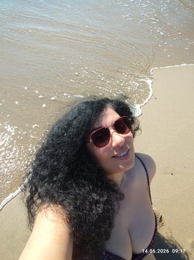 An overhead, close-up shot shows a woman with curly black hair and red-rimmed sunglasses on a sandy beach in Rezovo, Bulgaria. The woman is smiling, and her shoulders and chest are visible. She is wearing a dark bikini with a floral pattern. The water from the sea laps at the sandy shore. A timestamp in the bottom right corner of the image reads "14.06.2026 09:17".