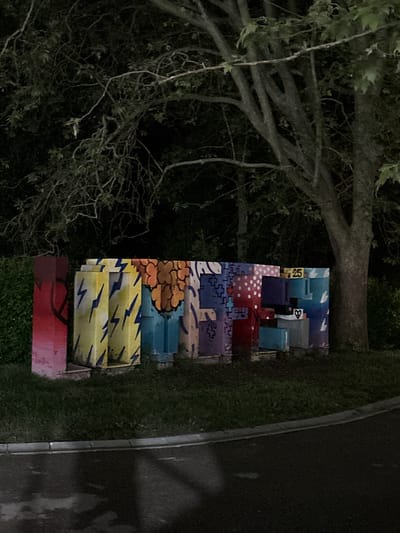 The image shows a colorful, art installation next to a dark asphalt road or walkway, possibly in Vrnjacka Banja Municipality, Serbia, though this cannot be forensically confirmed solely from the image. The installation appears to be constructed from painted concrete blocks or similar materials, arranged in a vertical stack. Each block features a distinct and varied design, including geometric patterns, lightning bolts, cloud-like shapes, and architectural motifs. The lighting is poor, suggesting nighttime or a dimly lit location. A tree with dense branches and foliage looms over the art installation on the left side of the frame. Green grass surrounds the base of the installation and lines the edge of the road. There are no people visible in the image. The composition is slightly rotated clockwise.