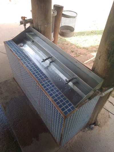 This is a photograph of a tiled, outdoor water trough with two metal spigots and a drain, situated next to a rough-hewn wooden post. A wire mesh trash can is also visible attached to the post behind the trough. The trough itself appears to be made of stainless steel and has a few water droplets on its surface. The tiling is a pattern of small, dark blue squares. The ground around the trough is concrete, and there is dry grass and dirt in the background. The overall impression is of a utilitarian public facility, possibly a drinking fountain or wash station, located in an outdoor setting. Given the context of Cuiabá, Brazil, this could be found in a park, public square, or recreational area.