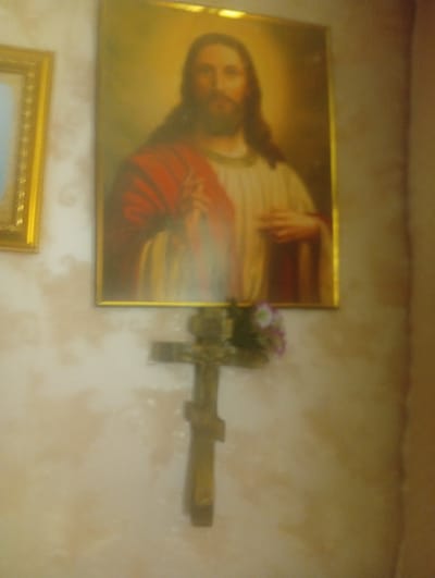 The image depicts a dimly lit interior scene with religious iconography. On the left, a framed picture of Jesus Christ is prominently displayed. The painting features Jesus with a beard and long hair, wearing a red garment over a white tunic, and his hands are clasped. Below the painting, a wooden cross is visible, adorned with a small bouquet of flowers. To the left of the Jesus painting, a sliver of another framed artwork can be seen. The background consists of a textured, light-colored wall, possibly wallpaper. The overall impression is of a domestic or religious space decorated with devotional items. There is no visible indication of the specific location within the city of Пліскі сельскі Савет, Belarus, beyond the general context of an indoor environment.