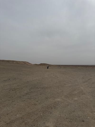 The image captures a vast, desolate landscape in Guazhou County, China. The scene is split between a grayish, overcast sky on the left and a brown, gravelly terrain on the right. A small, dark, human figure stands on the edge of a low escarpment or ridge that separates the sky from the terrain, appearing diminutive against the expansive background. The ground is littered with small stones and appears barren, with faint tracks suggesting previous passage. The horizon is barely visible, merging with the sky.
