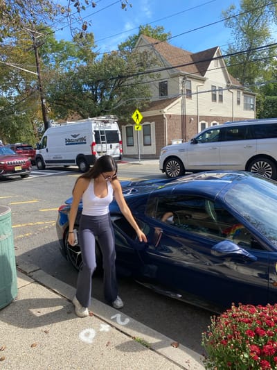 A young adult female is positioned next to a dark blue Ferrari sports car, its driver's door open, on a sidewalk in Ridgewood, United States. She wears a white halter top, dark gray flared leggings, white athletic shoes, and sunglasses, with a smartphone held in her left hand. A multi-colored accessory is visible inside the Ferrari through the passenger window. The immediate foreground features a green public waste bin on the left and a planter containing red chrysanthemums on the right, with white chalk markings on the concrete sidewalk. On the asphalt street, a white commercial van displaying "DREWING COMMERCIAL SERVICES" branding and a roof rack is parked, partially obscuring a red SUV. A white Lincoln Navigator SUV is also visible further down the street. Across the street stands a two-story building with light-colored siding, a brown roof, and a stone-clad lower level, with a yellow pedestrian crossing sign erected nearby. Overhead power lines and deciduous trees with green and autumn-tinted leaves are present against a blue sky.