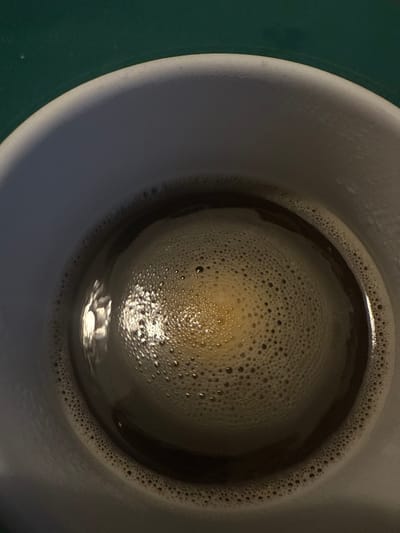 The scene, located in Arthès, France, presents a top-down view into a white or light grey ceramic mug. The mug contains a dark brown liquid, consistent with coffee, which is largely covered by a layer of fine, off-white froth. The froth displays a dense aggregation of small bubbles, arranged in a circular, somewhat concentric pattern across the liquid surface, with a slight visual density towards the center. A prominent, elongated reflection of an external light source, appearing bright white, is visible on the upper-left quadrant of the liquid, interacting with the froth and the underlying liquid. The upper inner rim of the mug is visible, and the mug rests on a dark green surface, portions of which are discernible at the top and top-left edges of the image. No individuals, specific actions, or interactions are depicted.